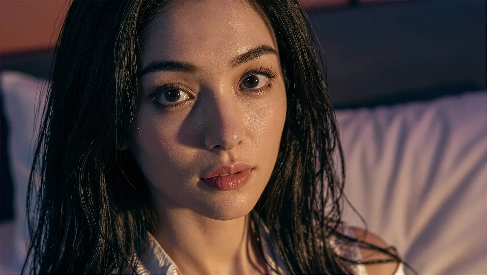 Fictional AI character lying on a pillow in warm lamplight, looking straight into the lens